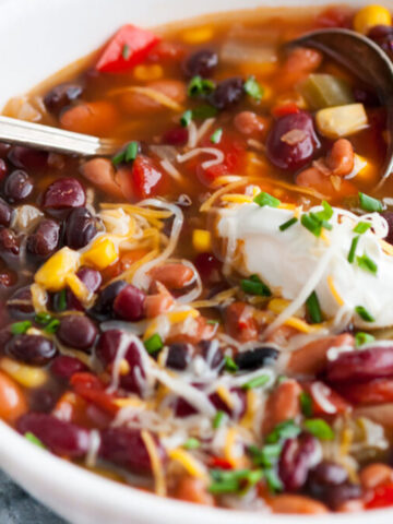 Slow Cooker Vegetarian Three Bean Chili in white bowl on gray marble with silver spoon, melted cheese and dollop of sour cream close up view