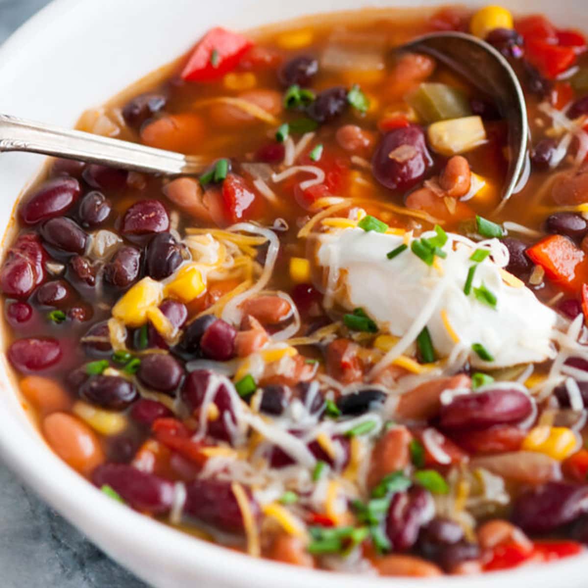 Slow Cooker Vegetarian Three Bean Chili in white bowl on gray marble with silver spoon, melted cheese and dollop of sour cream close up view