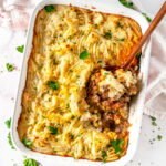Shepherd's Pie in white casserole dish on white marble with spoonful of filling in wooden spoon overhead view