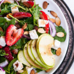 Strawberry Spinach Arugula Salad on white plate and gray platter with sliced apples and berries overhead view