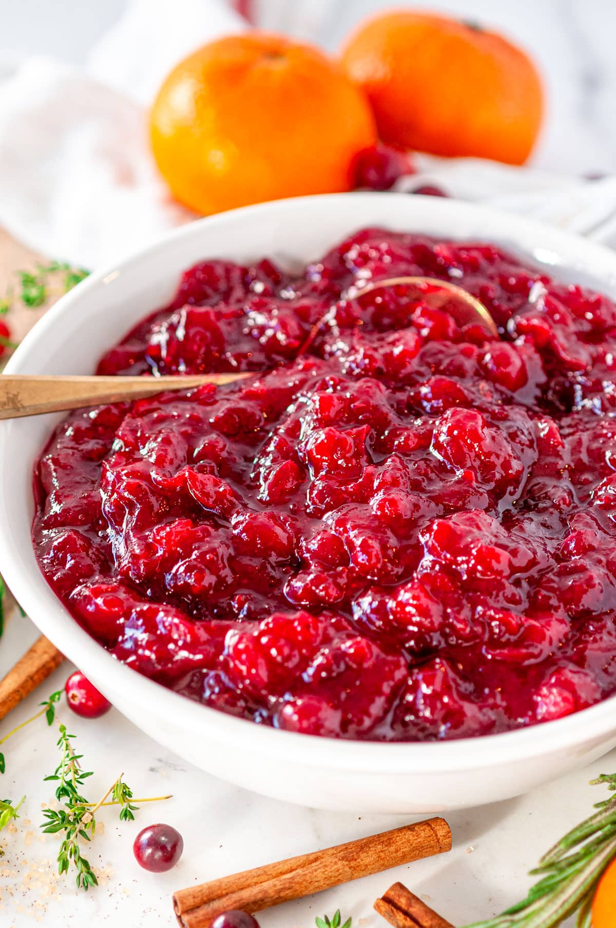 Holiday Orange Cranberry Sauce in white bowl with gold spoon, fresh rosemary, thyme, oranges, and cinnamon sticks on white marble