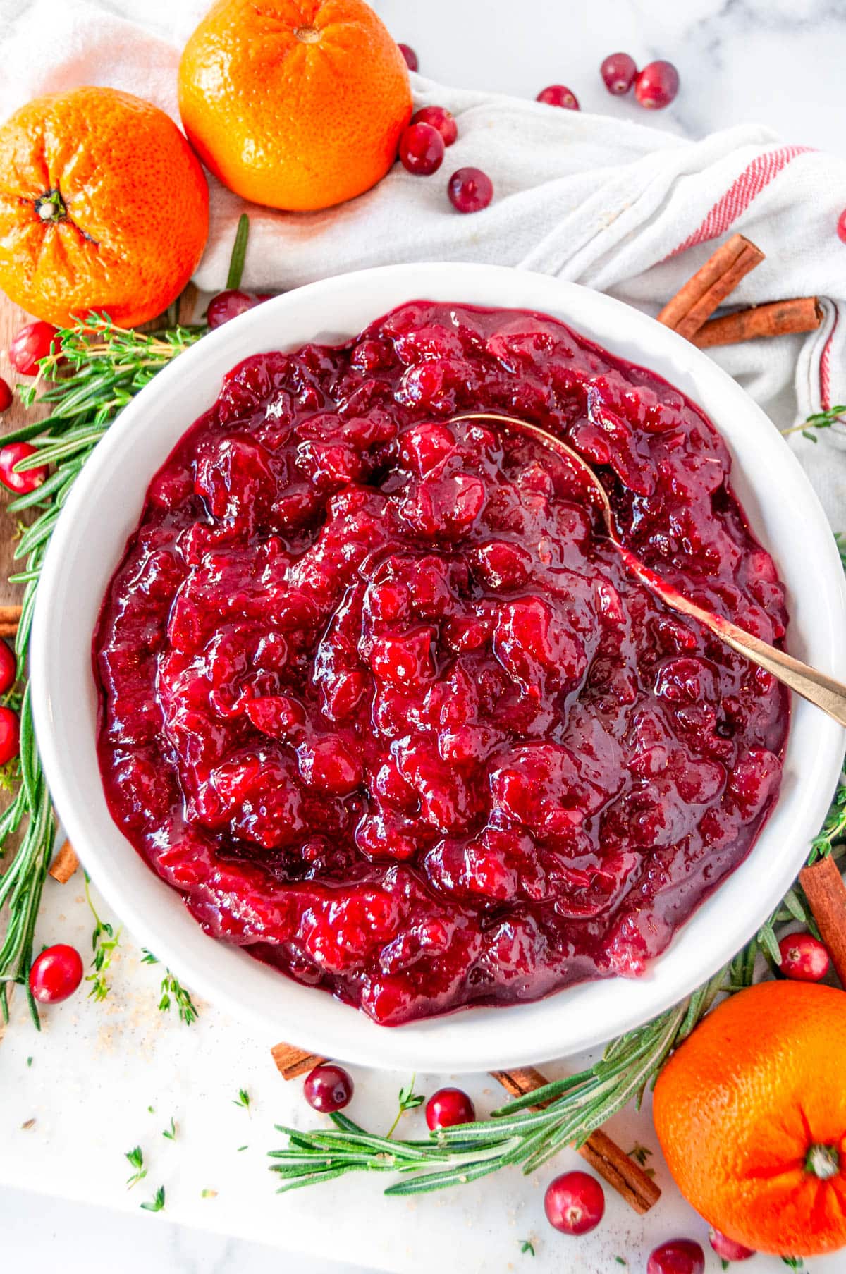 Holiday Orange Cranberry Sauce in white bowl with gold spoon, fresh rosemary, thyme, oranges, and cinnamon sticks on white marble