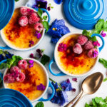 Limoncello Creme Brulee in blue le creuset cocottes with raspberries, flowers, mint leaves and gold spoons on white marble