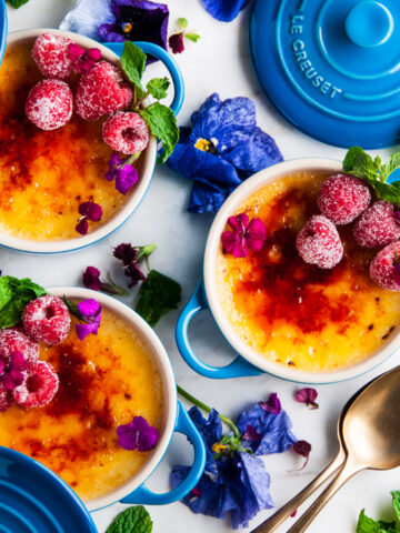 Limoncello Creme Brulee in blue le creuset cocottes with raspberries, flowers, mint leaves and gold spoons on white marble