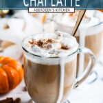 Pumpkin Spice Chai Latte in glass mugs with whipped cream on and gold spoon on white marble - aqua rectangle with white text overlay