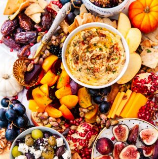 Greek Autumn Cheese Board on white marble with hummus, gold and red beets, honey drizzled figs and dates, olives and feta cheese, grapes, apples, cheddar cheese, pomegranate seeds, pistachios, nuts, cranberries and pita chips