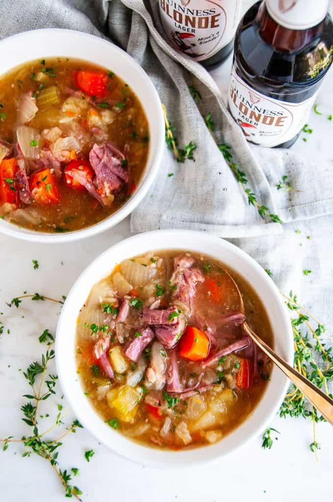Slow Cooker Corned Beef and Cabbage Stew in white bowls with gold spoon, gray tea towel, and Guinness blond lager