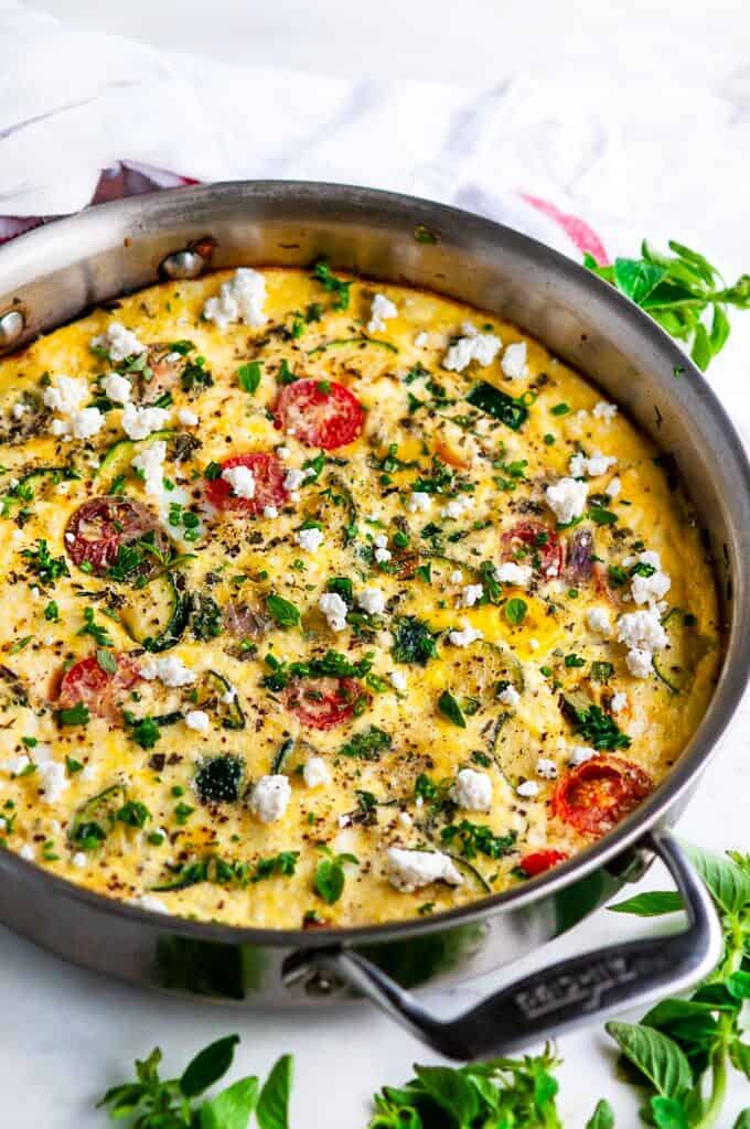 Greek Vegetable Frittata in all-clad skillet with oregano and tea towel on white marble