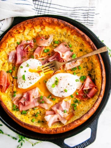 Savory Herb Cheddar Dutch Baby with poached eggs and prosciutto in lodge cast iron skillet with gold fork and tea towel on white marble