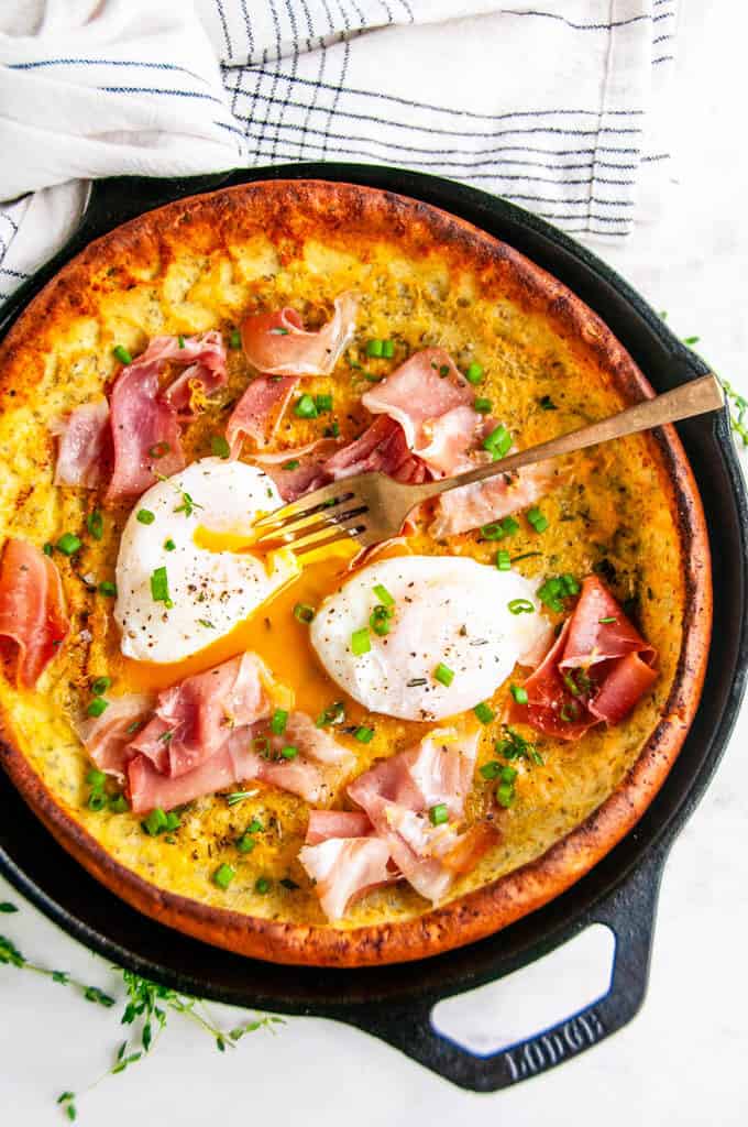 Savory Herb Cheddar Dutch Baby with poached eggs and prosciutto in lodge cast iron skillet with gold fork and tea towel on white marble