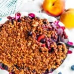 Easy Peach Blueberry Crisp in white pie pan with tea towel and serving spoon on white marble