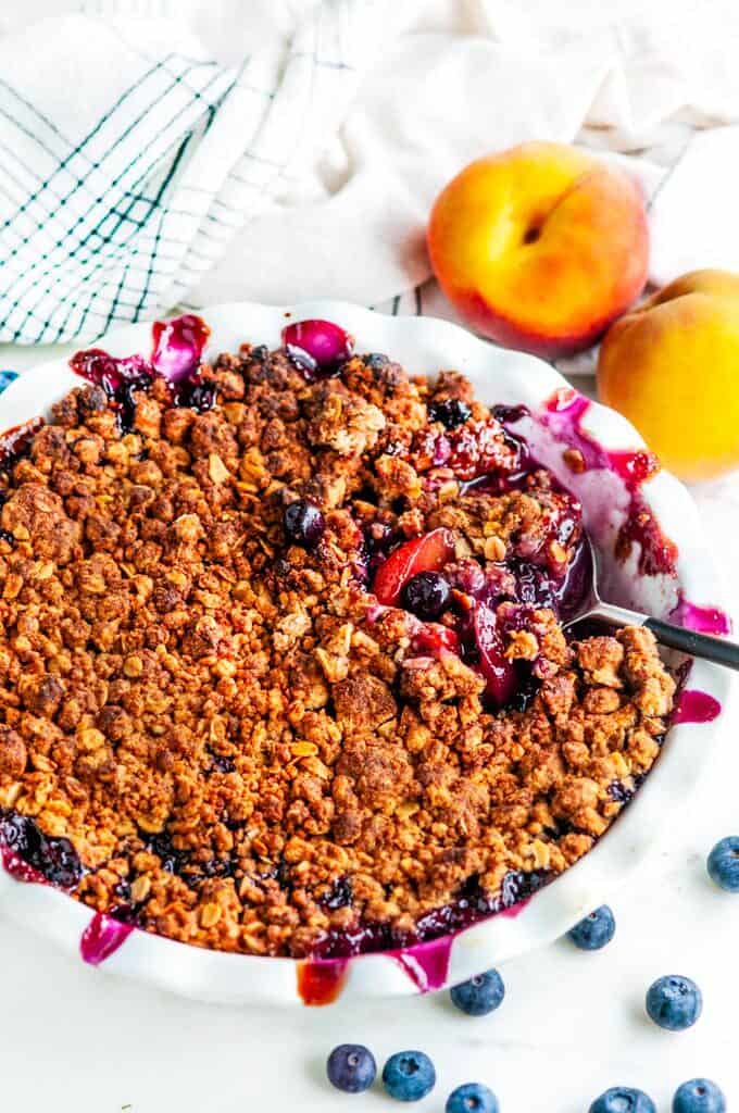 Easy Peach Blueberry Crisp in white pie pan with tea towel and serving spoon on white marble