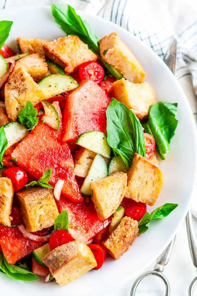 Watermelon Panzanella Salad in white bowl with silver serving tongs and tea towel close up