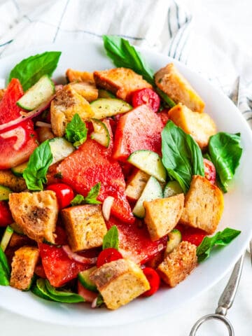 Watermelon Panzanella Salad in white bowl with silver serving tongs and tea towel