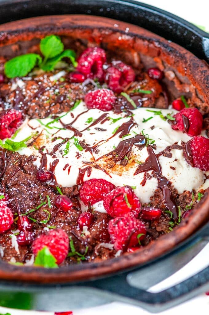 Chocolate Dutch Baby with whipped cream, chocolate sauce, rasperries, mint, pomegranate arils, and powdered sugar in lodge cast iron skillet on white marble close up