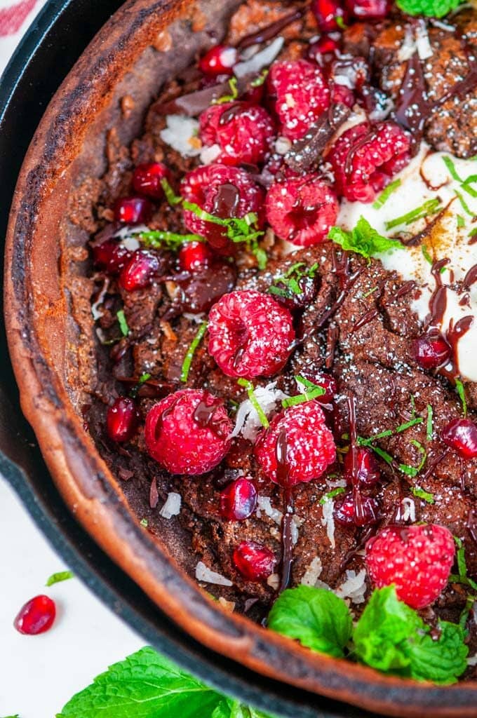 Chocolate Dutch Baby with whipped cream, chocolate sauce, rasperries, mint, pomegranate arils, and powdered sugar in lodge cast iron skillet on white marble close up