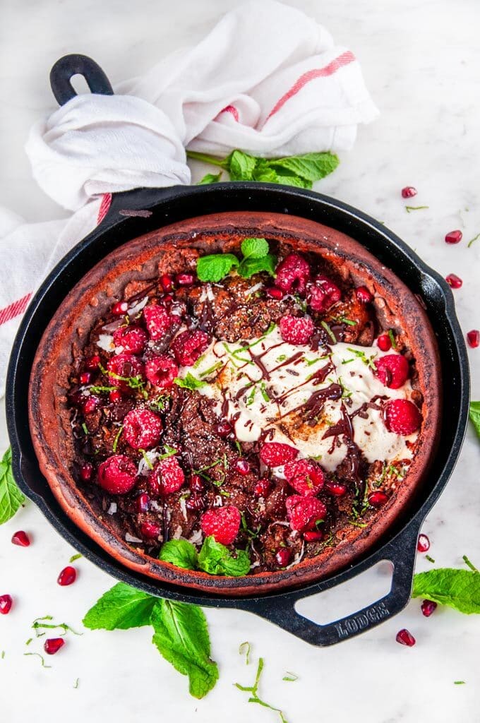 Chocolate Dutch Baby with whipped cream, chocolate sauce, rasperries, mint, pomegranate arils, and powdered sugar in lodge cast iron skillet on white marble
