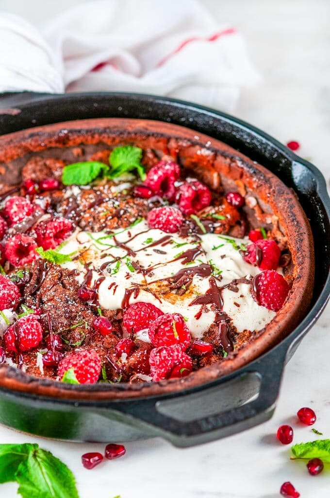 Chocolate Dutch Baby with whipped cream, chocolate sauce, rasperries, mint, pomegranate arils, and powdered sugar in lodge cast iron skillet on white marble