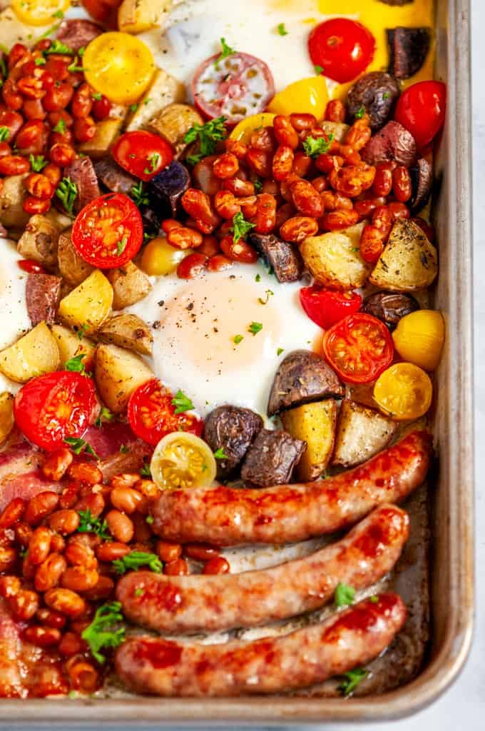 Sheet Pan Full English Breakfast with sausage, bacon, eggs, tomatoes, baked beans, and potatoes on white marble close up