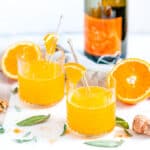 Immune Boosting Orange Turmeric Cocktail in glasses with fresh sage, prosecco bottle, and tea towel on white marble