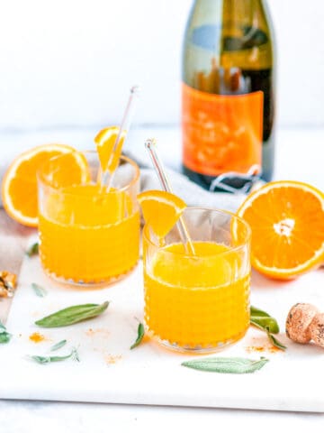 Immune Boosting Orange Turmeric Cocktail in glasses with fresh sage, prosecco bottle, and tea towel on white marble