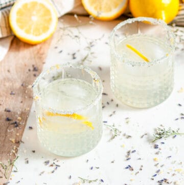 Lavender Collins Cocktail with lemons and soda bottles on white marble