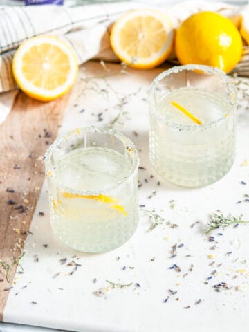 Lavender Collins Cocktail with lemons and soda bottles on white marble