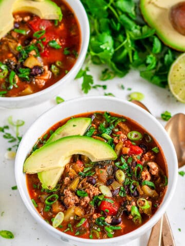 One Pot Turkey Chili in white bowls with gold spoons, avocado, cilantro and limes on marble