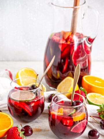 Fruity Red Wine Sangria with citrus fruits and cherries in pitcher and stemless wine glasses with gold spoons