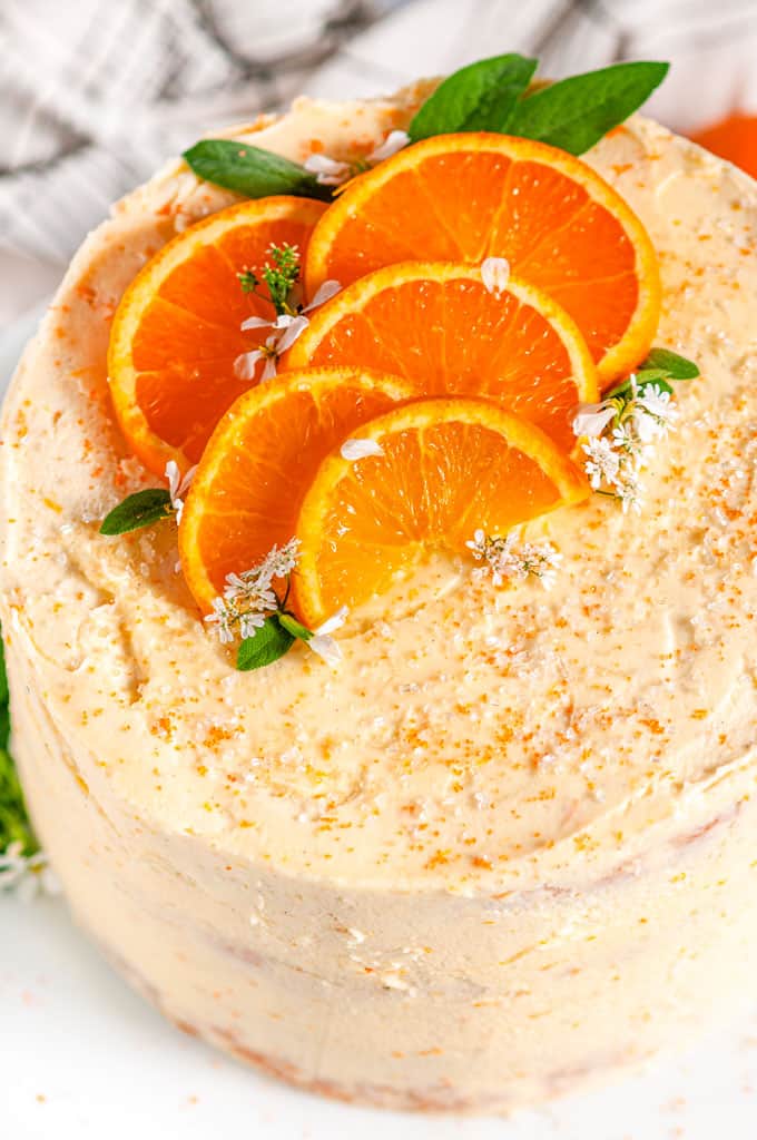 Orange Cake with Zesty Cream Cheese Frosting with orange slices and sage leaves on white cake stand over head close up