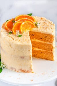 Orange Cake with Zesty Cream Cheese Frosting with orange slices and sage leaves on white cake stand slice removed side view