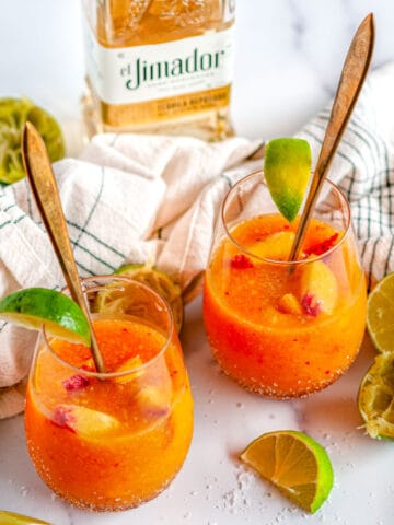 Frozen Peach Margaritas in glasses with lime slices and gold stirrers on white marble with tea towel and tequila bottle in background