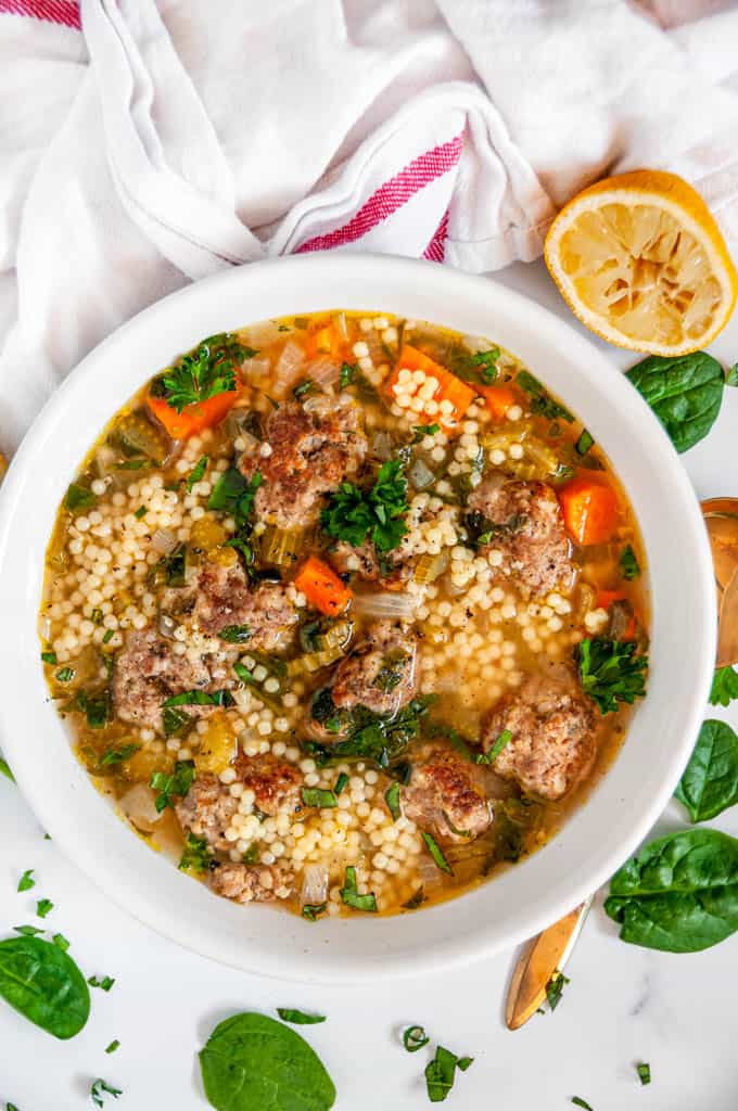 Italian Wedding Soup in white bowl with gold spoon, spinach, and a squeezed lemon 