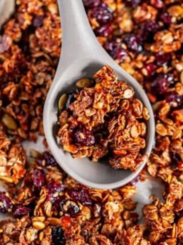 Chai Spice Cranberry Pecan Granola on sheet pan with gray silicone wooden spoon held in hand