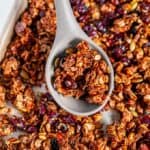 Chai Spice Cranberry Pecan Granola on sheet pan with gray silicone wooden spoon held in hand