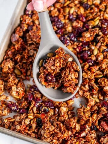 Chai Spice Cranberry Pecan Granola on sheet pan with gray silicone wooden spoon held in hand