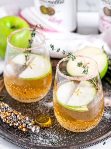 Sparkling Apple Cider Gin Cocktail in two glasses with jingle bell bottle opener on gray plate