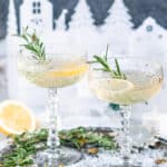 Sparkling Elderflower Gin Cocktail in crystal glasses with rosemary, lemon slices, and paper holiday village in background