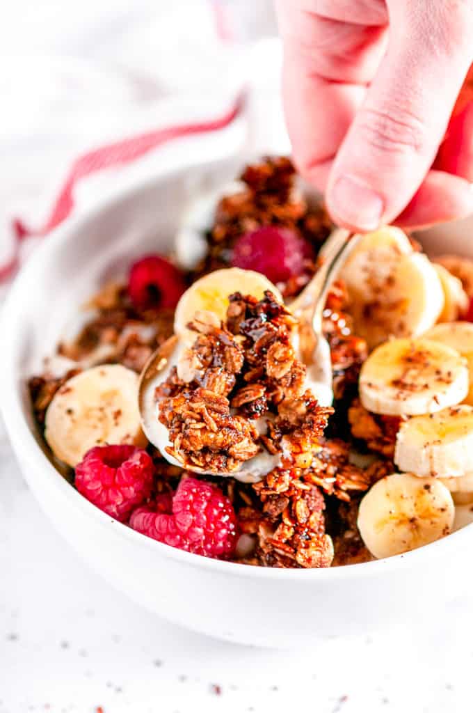 Crunchy Walnut Banana Bread Granola in white bowl with raspberries, sliced bananas, and gold spoon held in hand