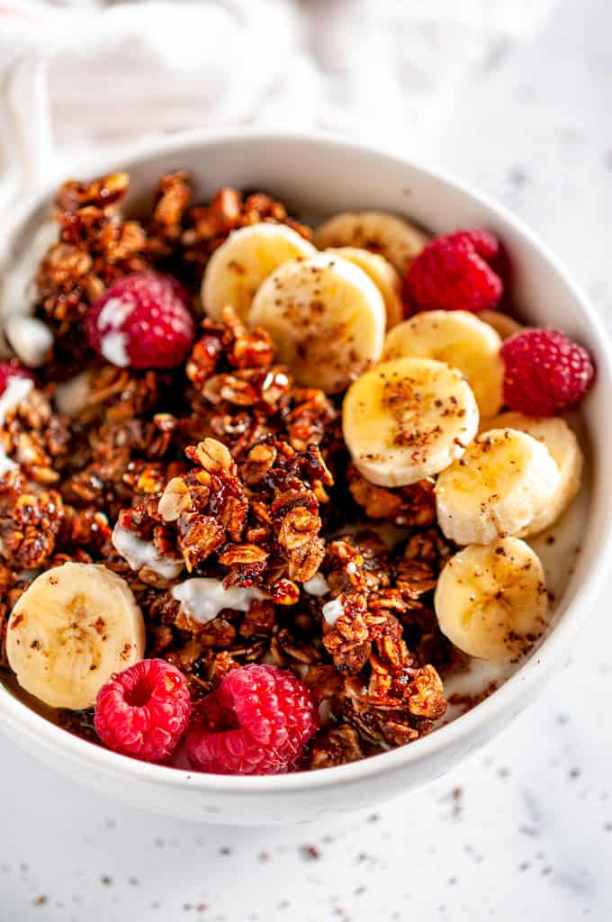 Crunchy Walnut Banana Bread Granola in white bowl with raspberries and sliced bananas on white marble