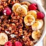 Crunchy Walnut Banana Bread Granola in white bowl with raspberries, sliced bananas, and gold spoon