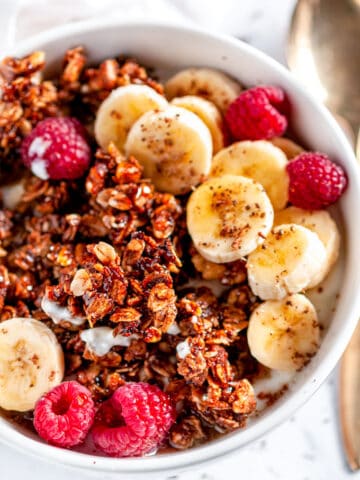 Crunchy Walnut Banana Bread Granola in white bowl with raspberries, sliced bananas, and gold spoon
