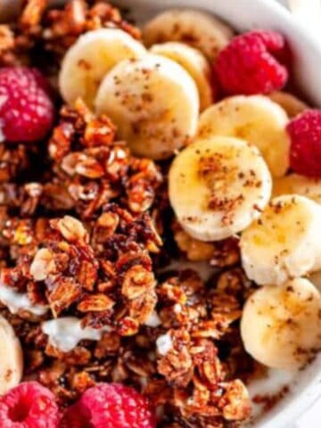 Crunchy Walnut Banana Bread Granola in white bowl with raspberries, sliced bananas, and gold spoon