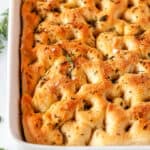 Easy Herbed Focaccia Bread in white baking dish with rosemary and oregano on white marble