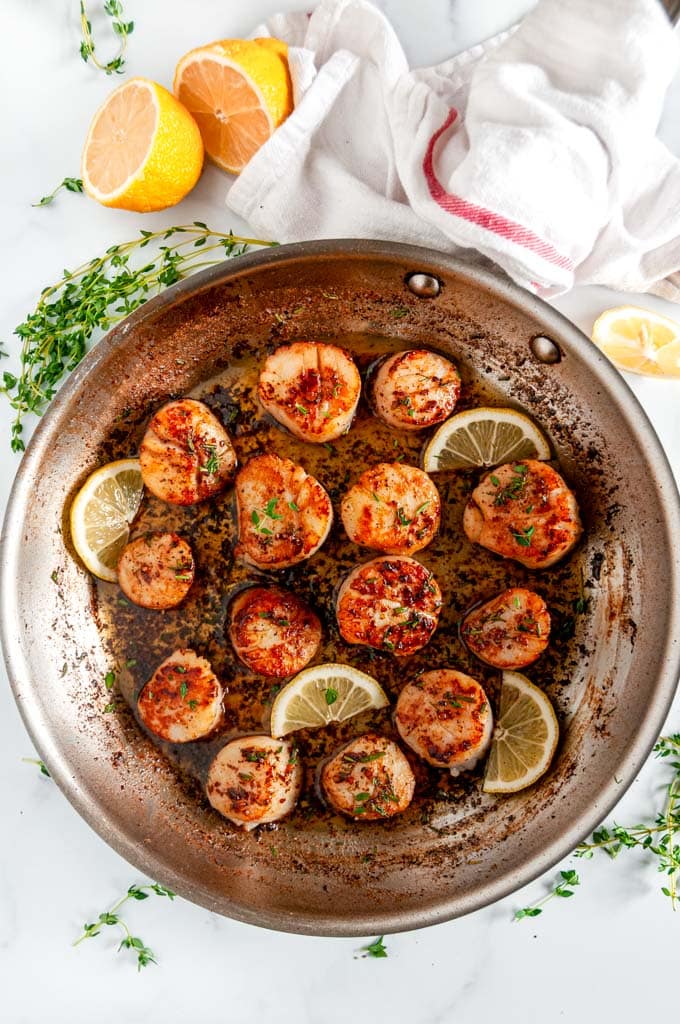 Pan Seared Lemon Garlic Butter Scallops in all-clad skillet with lemon slices and herbs on white marble