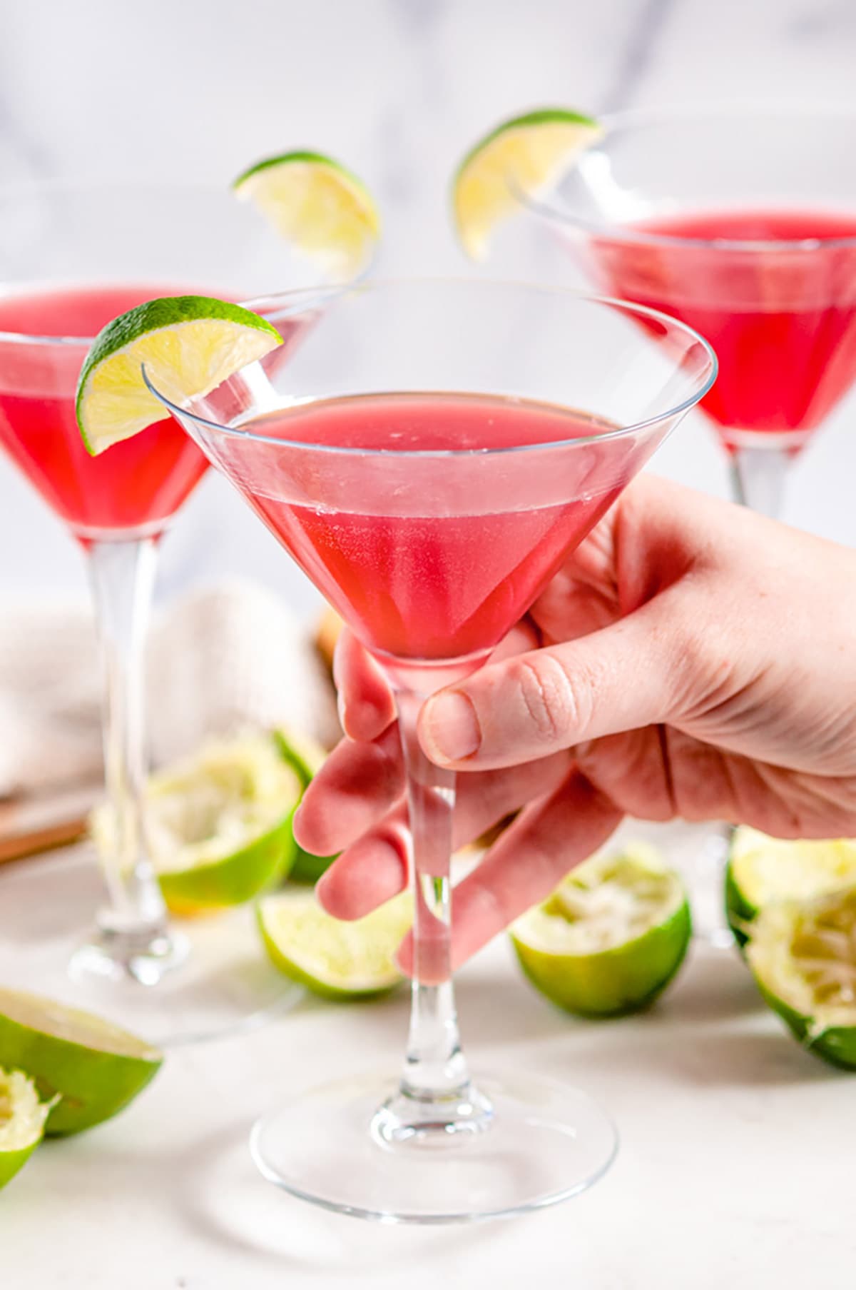 Classic Cosmopolitan Cocktail in martini glasses with lime wedge held in hand on white marble
