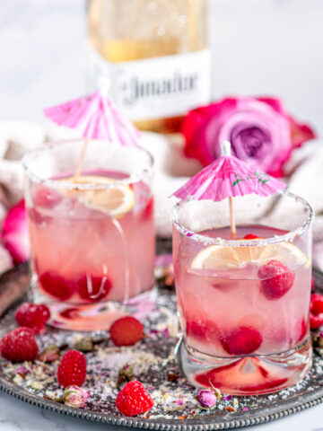 Sparkling Raspberry Senorita Margarita in rose sugar rimmed glasses with pink umbrellas on a gray plate, El Jimador tequila bottle in background