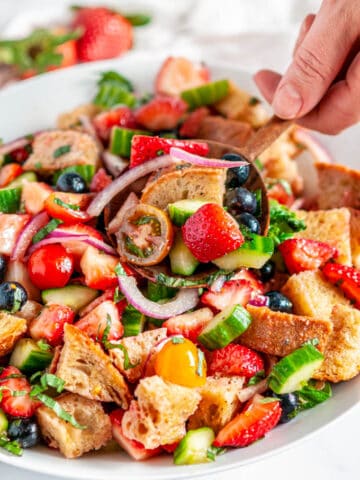 Berry Tomato Panzanella Salad in white bowl on marble with wooden spoon held in hand