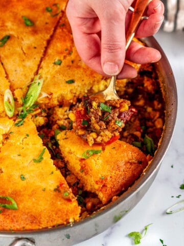 Homemade Cornbread Tamale Pie in All-Clad skillet with gold spoon held in hand on white marble close up