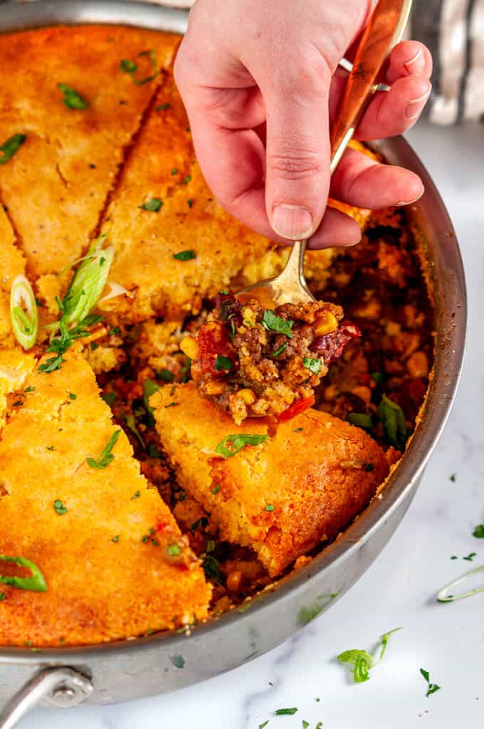 Homemade Cornbread Tamale Pie in All-Clad skillet with gold spoon held in hand on white marble close up
